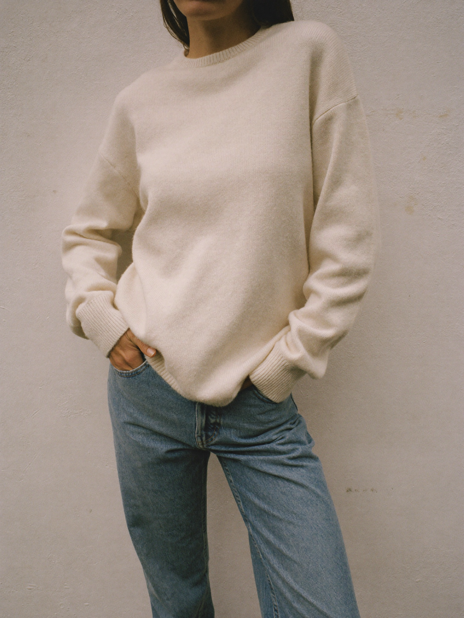 A plain oatmeal crewneck knit sweater and denim against a warm ivory plaster wall