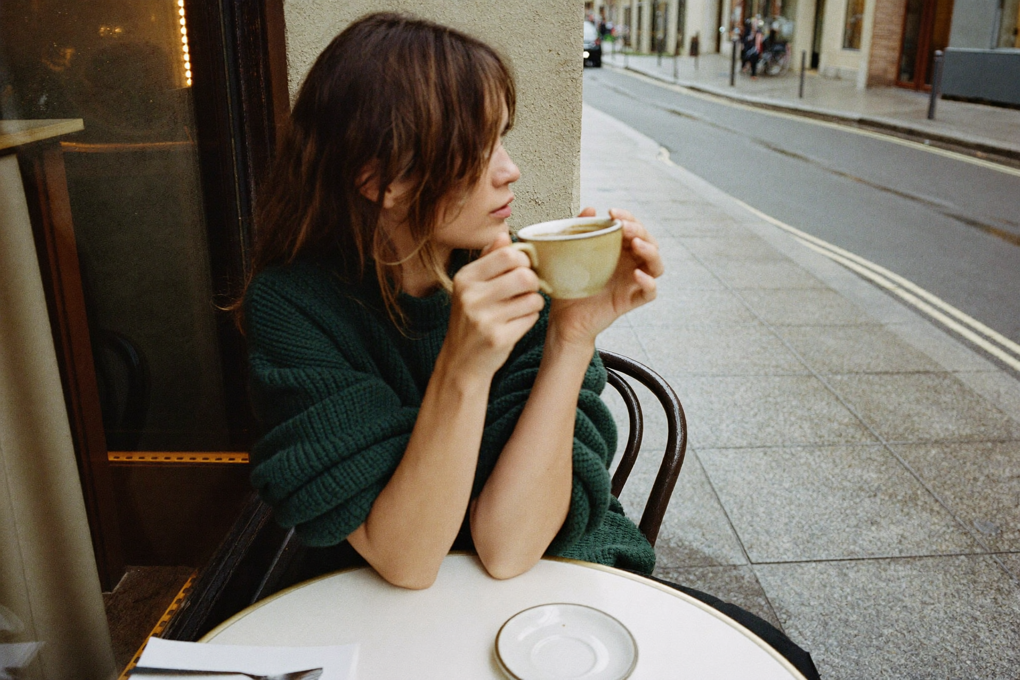 Knit and Loom editorial break desktop - a woman in a deep green chunky knit sweater at a cafe terrace holding a stoneware cup