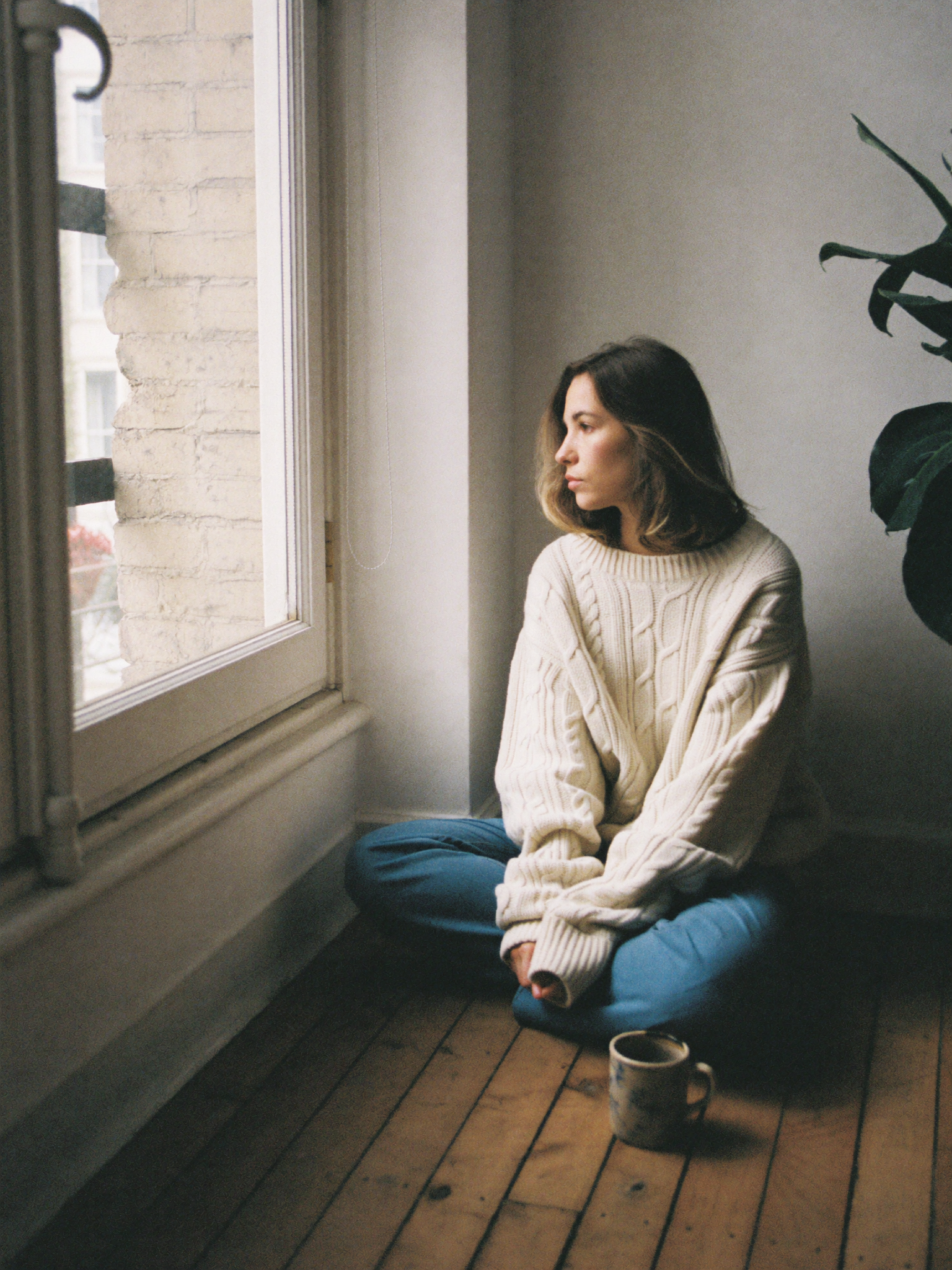 Knit and Loom brand section - a woman in an ecru cable knit sweater sitting on a wooden floor beside a tall window